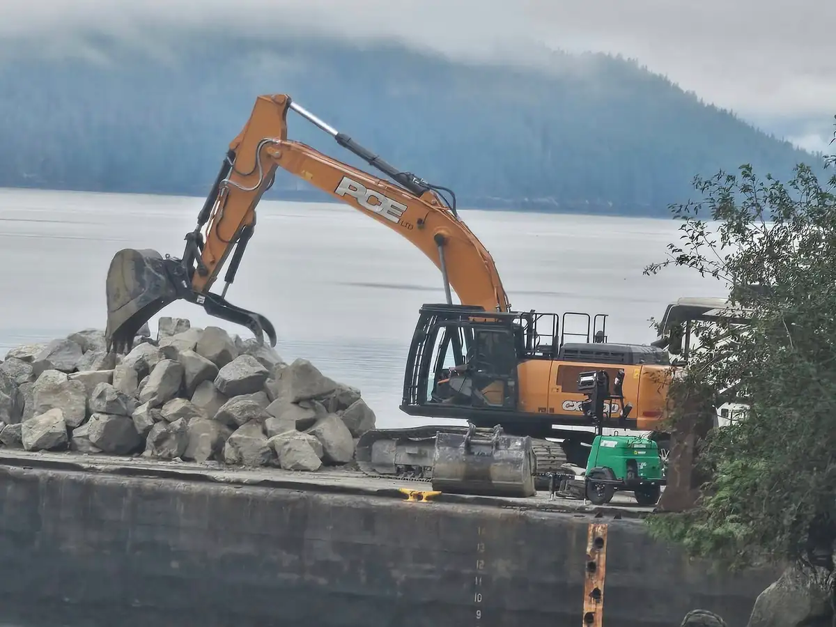 PCE excavator working rock on a barge at Rivers Inlet, mountains behind