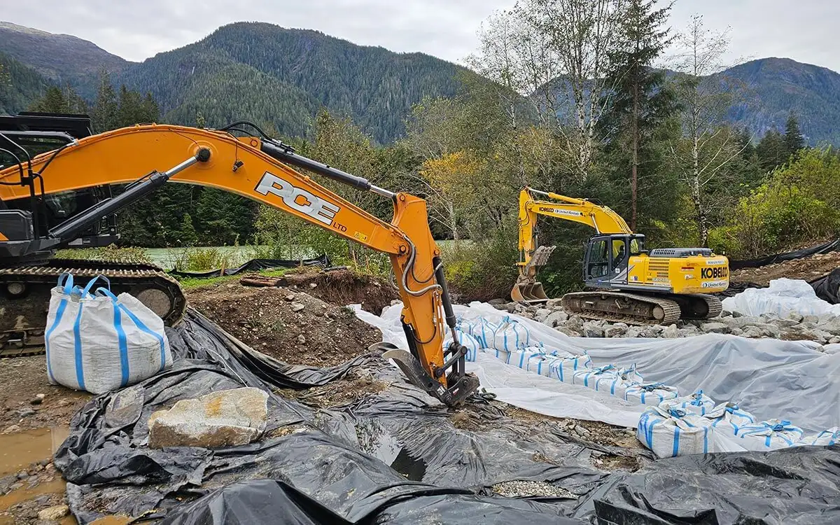 Excavator working beside poly-covered stockpiles near the creek