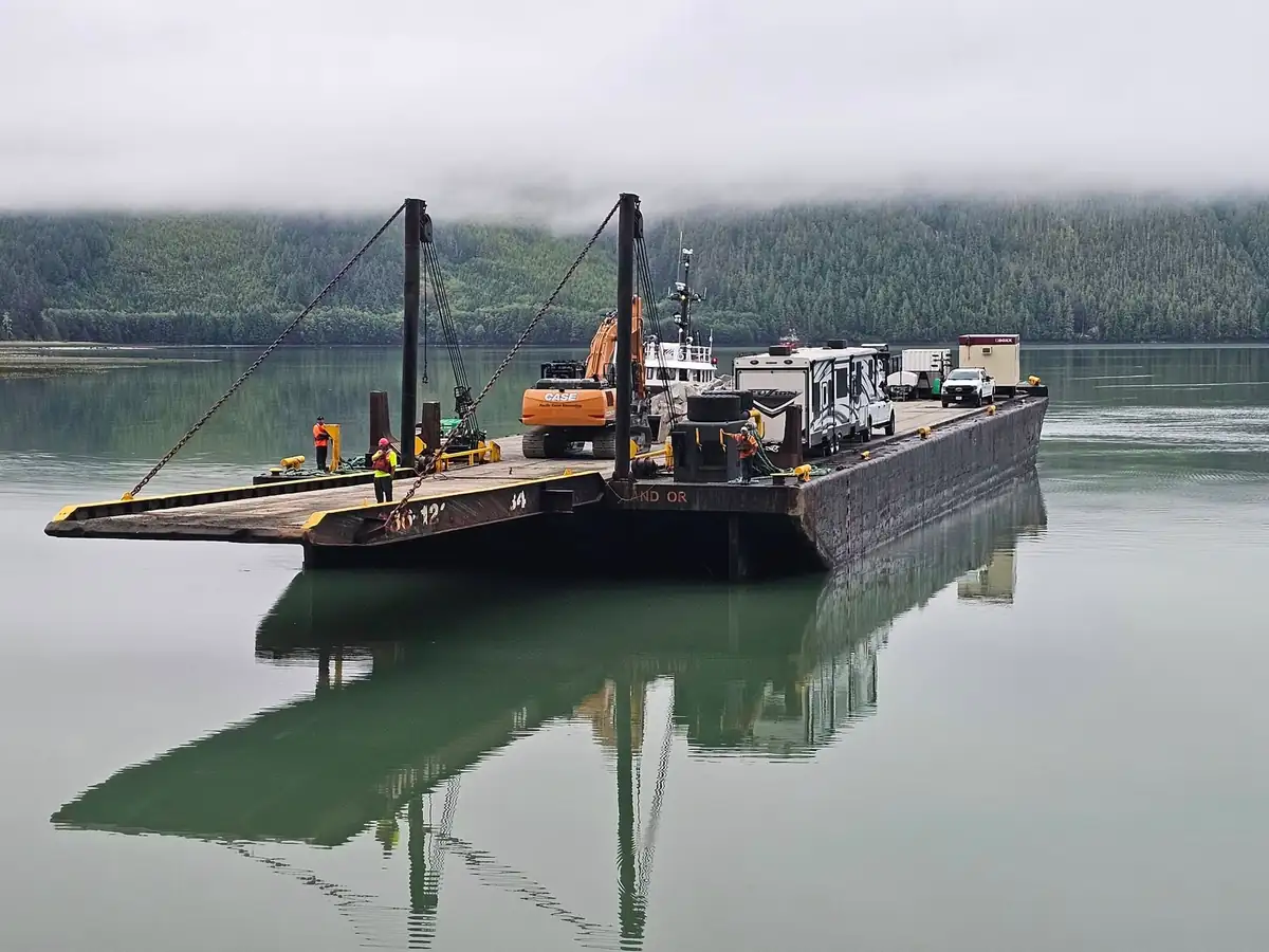 Tug and loaded barge carrying excavators and trailers on a calm inlet