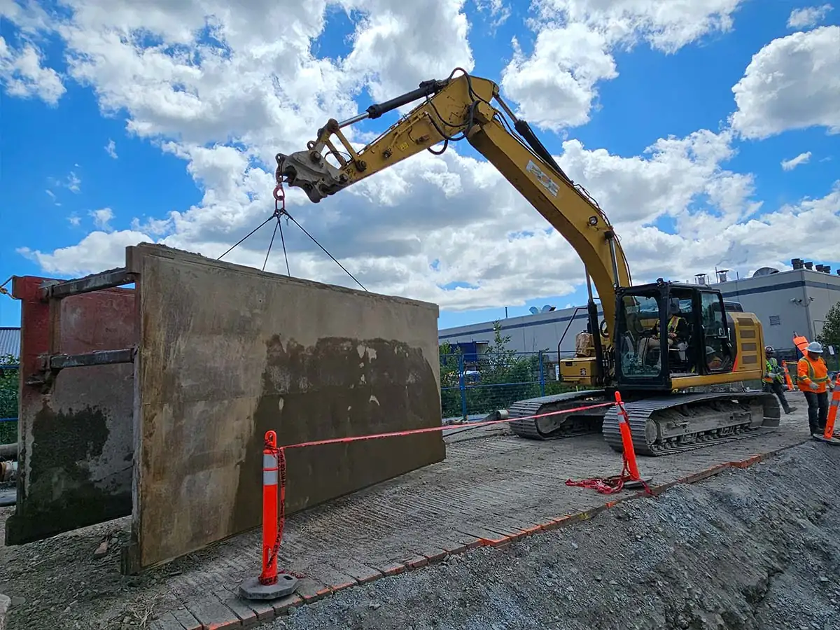 Excavator lifting a steel shoring panel beside a trench
